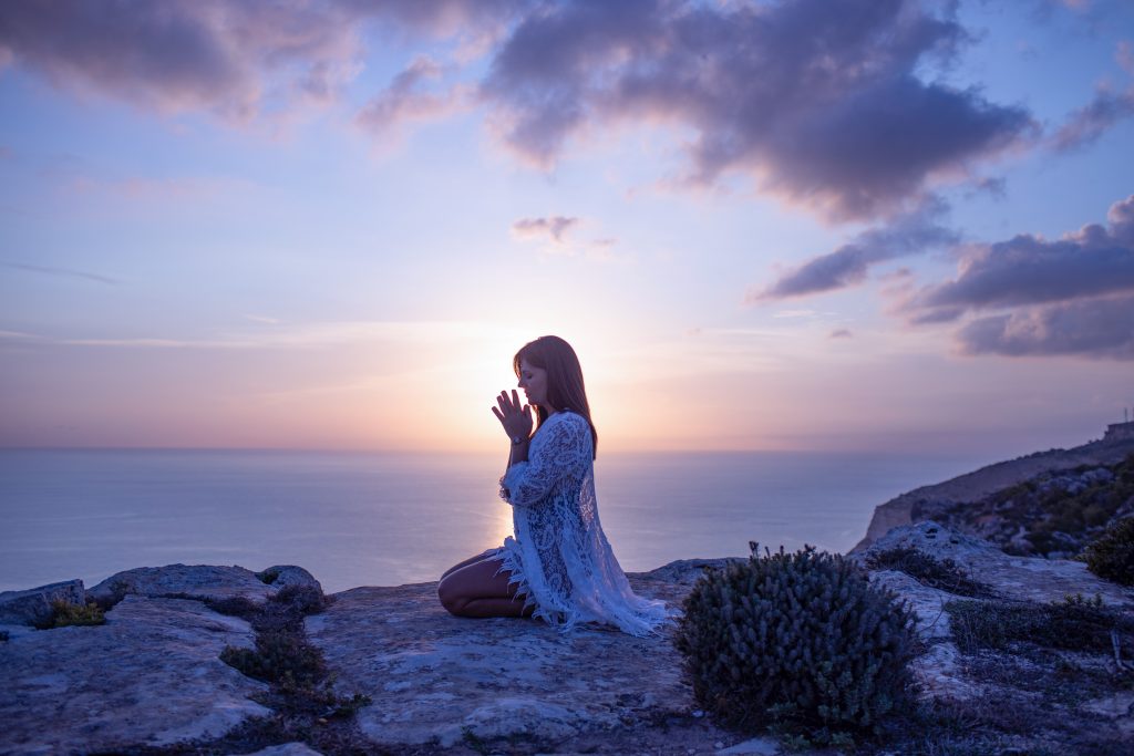 Woman connecting to herself through embodiment with her hands in a praying pose on a cliff.