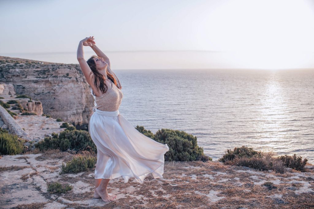 Woman with her arms up, practicing embodiment on a cliff with a sea view
